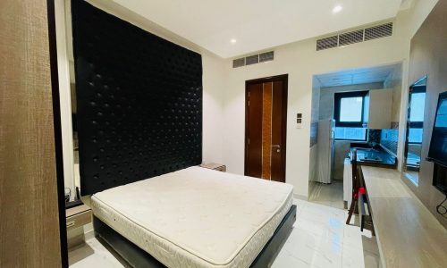 Luxury studio apartment with a large black headboard, white mattress, glossy white floor tiles, and a kitchenette visible in the background.