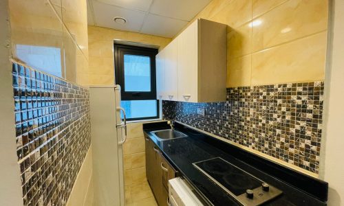 Luxury studio apartment kitchen with glass mosaic backsplash, brown cabinets, black countertop with built-in stove, and a fridge next to a narrow window.