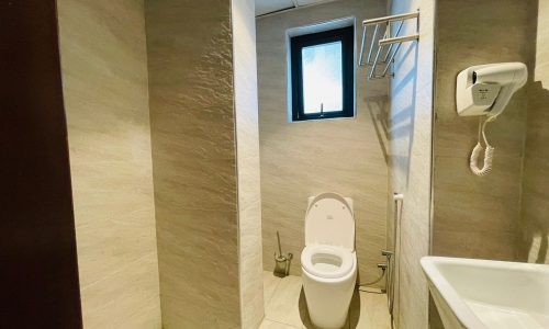 A small bathroom in a luxury studio apartment featuring a toilet, bidet, sink, and a shower area, with a towel rack and a small, open window on the upper wall.