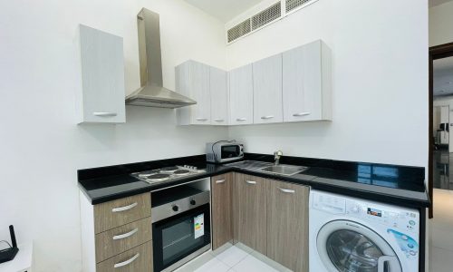 A modern kitchen in a luxury studio apartment with white cabinets, black countertops, built-in appliances including a stove, oven, and washing machine, and a stainless steel range hood.