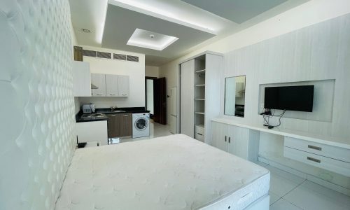 A small, modern studio apartment in Burhama with a bed in the foreground and an integrated kitchen area in the background, featuring white cabinets and modern appliances.