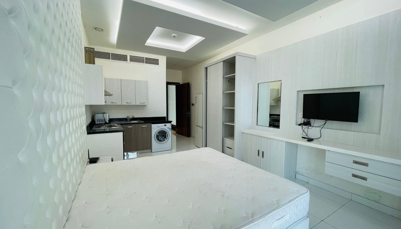 A small, modern studio apartment in Burhama with a bed in the foreground and an integrated kitchen area in the background, featuring white cabinets and modern appliances.
