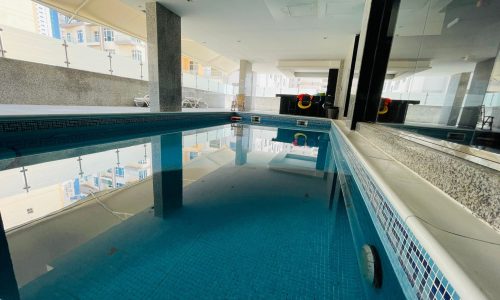 Indoor swimming pool with clear water, surrounded by white columns in a luxury studio apartment, overlooking buildings through large windows.