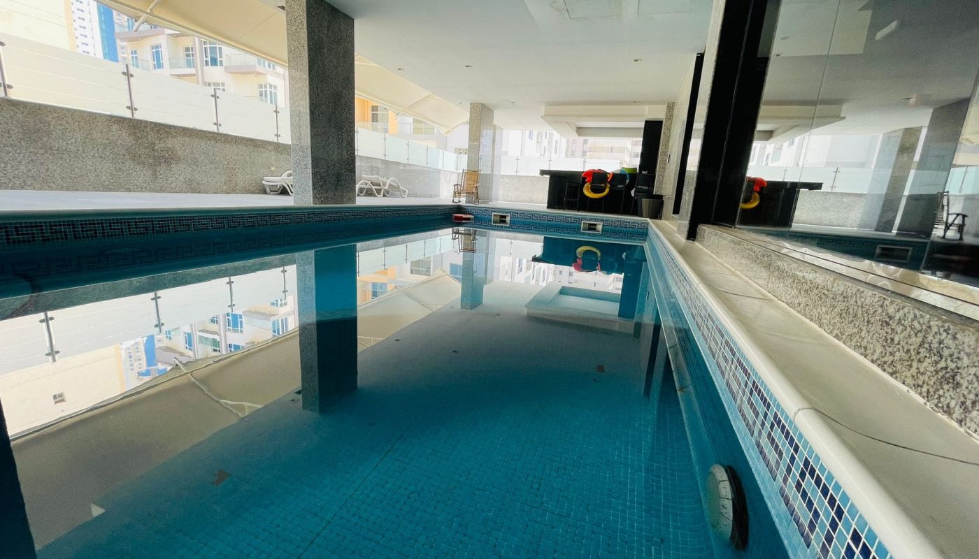 Indoor swimming pool with clear water, surrounded by white columns in a luxury studio apartment, overlooking buildings through large windows.