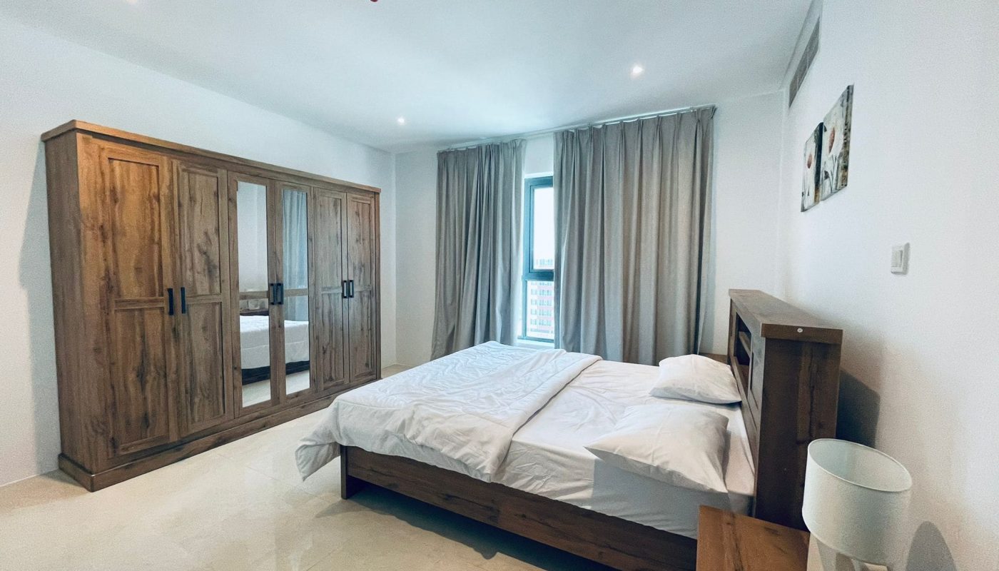 A luxury 1 BR apartment bedroom in Burhama, featuring a large bed, wooden furniture, and cream curtains on two windows.