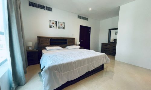 A modern, minimalist bedroom in a luxury 1 BR apartment featuring a double bed, wooden furniture, light gray walls, and a large window with curtains.