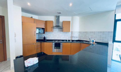 A modern kitchen in a luxury 2BR apartment with wooden cabinets, black countertops, and built-in appliances. There's a large window letting in natural light.