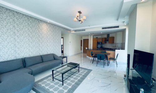 Modern living room in a luxury 2BR apartment with a gray sectional sofa, glass coffee table, patterned wall, and an open kitchen in the background.