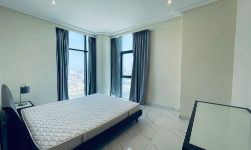 Modern, minimalist bedroom in a luxury 2BR apartment, featuring a large bed without bedding, grey curtains, and a city view from floor-to-ceiling windows.