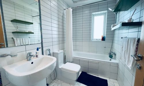 A clean, well-lit bathroom in a Luxury 2BR Apartment with a sink, toilet, and bathtub, featuring white tiles and a small window.