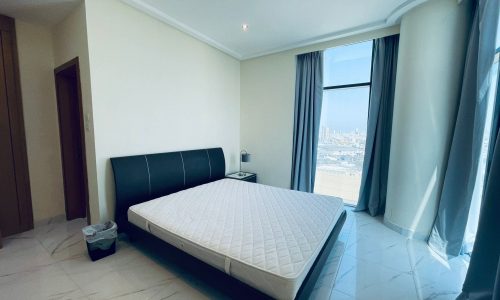 A modern bedroom in a luxury 2BR apartment, with a large bed, white mattress, teal curtains, and a city view of Sanabis through the window.