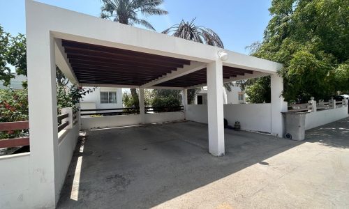 A semi-furnished villa with a carport shelter, featuring a white frame and wooden slat roof, surrounded by greenery and palm trees, under a clear sky.