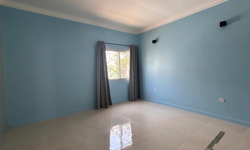 Empty room painted light blue in a semi-furnished villa, featuring a single window with closed gray curtains, reflecting sunlight on a white tiled floor.