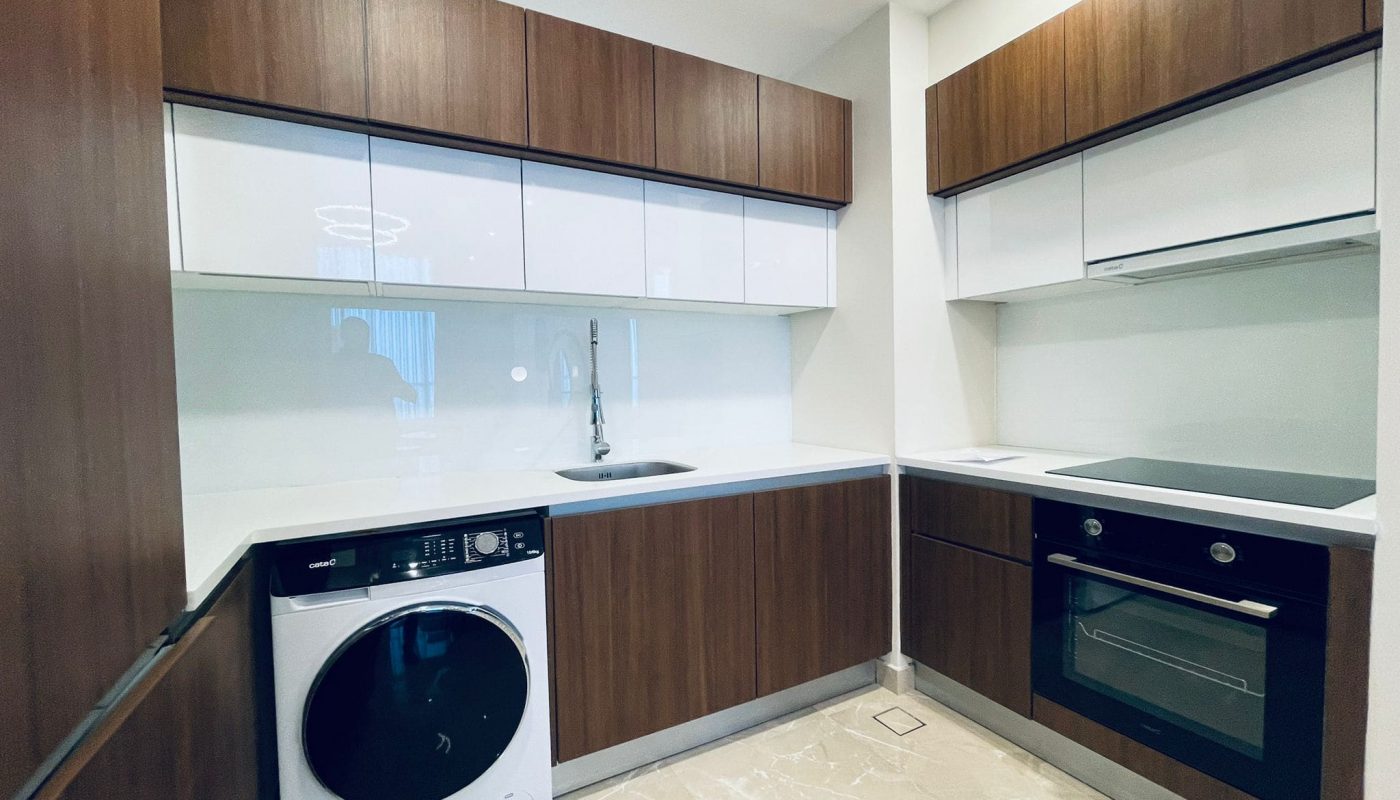 Modern kitchen interior featuring wooden cabinets, white countertops, built-in oven, washing machine, and reflective backsplash.