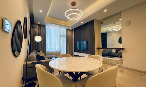 Modern living room with a marble dining table, beige chairs, and a sofa. features circular ceiling lights, wood paneling, and a mounted tv.