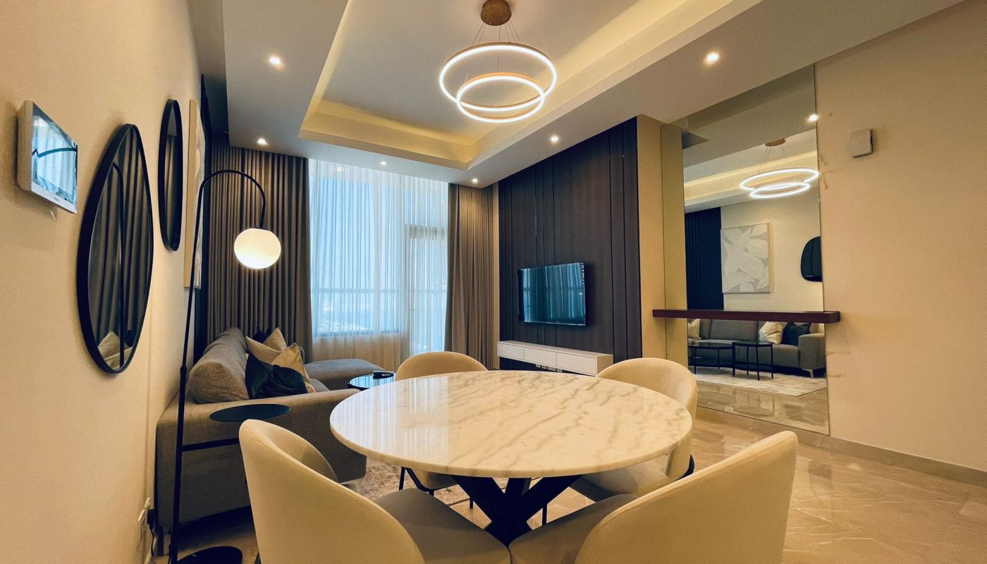 Modern living room with a marble dining table, beige chairs, and a sofa. features circular ceiling lights, wood paneling, and a mounted tv.