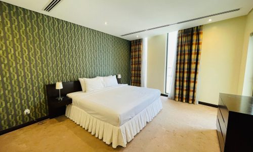 A modern luxury apartment in Sanabis with a large bed, patterned green wallpaper, striped curtains, and a minimalist desk.