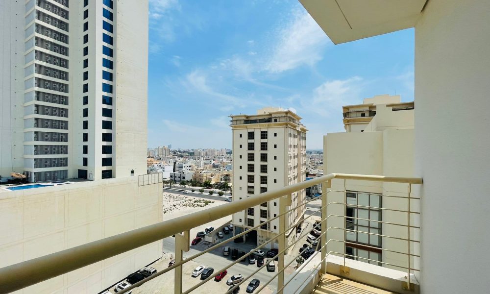 View from a luxury apartment balcony in Sanabis, overlooking other apartment buildings, a parking lot, and a distant urban landscape under a clear blue sky.