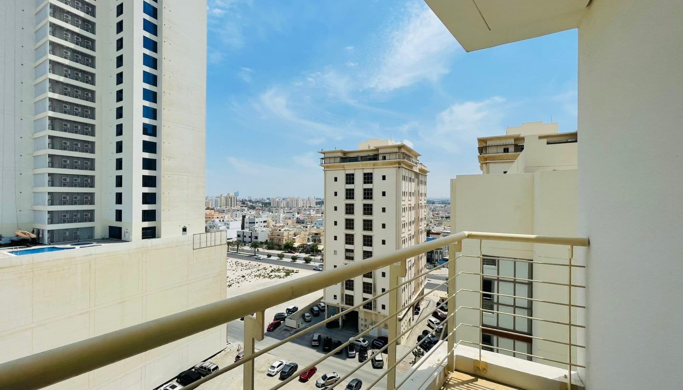 View from a luxury apartment balcony in Sanabis, overlooking other apartment buildings, a parking lot, and a distant urban landscape under a clear blue sky.