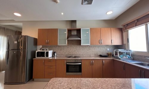 Modern kitchen interior with wooden cabinets, stainless steel refrigerator, gas stove, microwave, and a sink by a window.