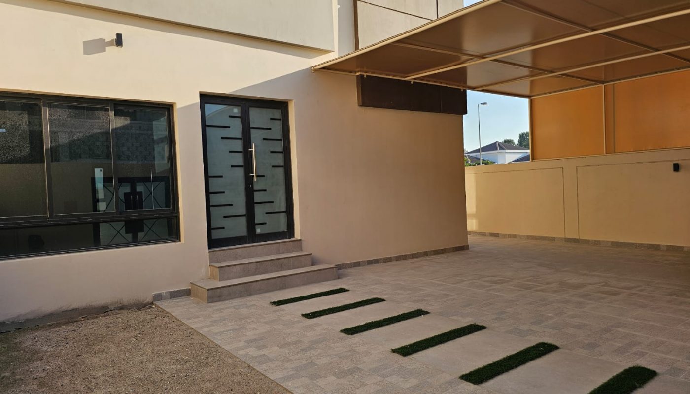 Modern house exterior with beige walls, a glass door with geometric patterns, tiled patio, covered parking area, and small green grass patches embedded in the ground.