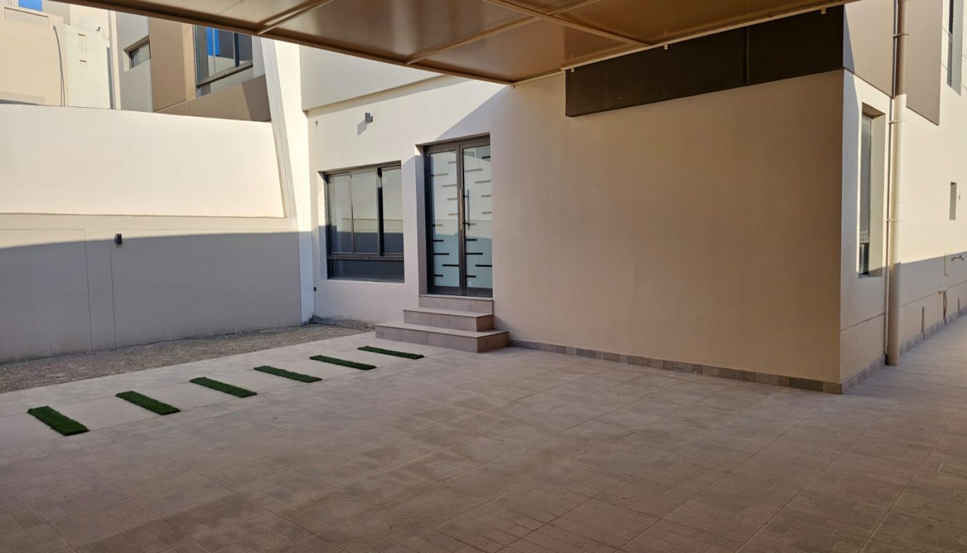 Covered outdoor area with tiled flooring, a few artificial grass strips, and a building entrance with frosted glass door and steps. Light-colored walls and windows are visible.