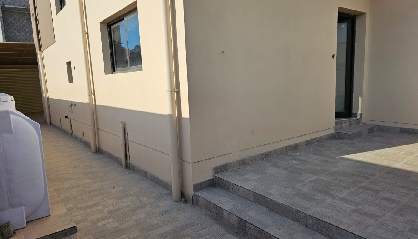 A beige building with a black-framed window and door, stone steps, and paved ground in bright daylight.