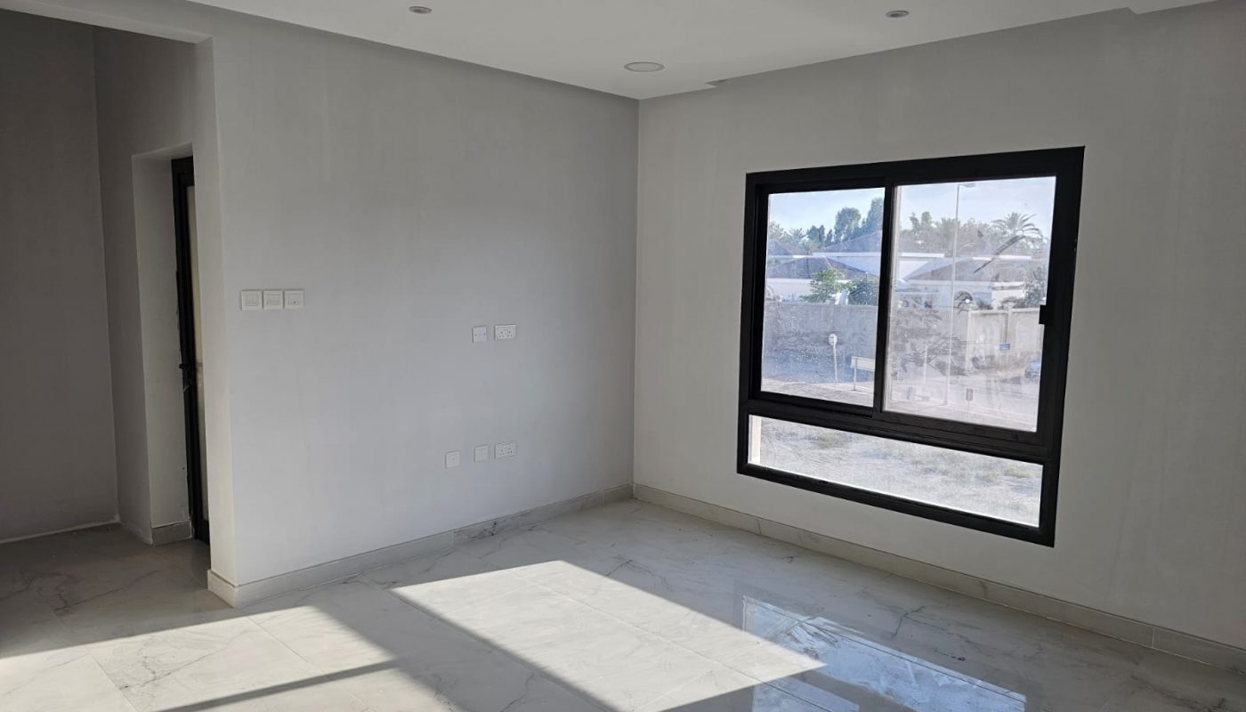 A bright, empty room with white walls, a large window, tiled floor, and electrical outlets on the wall. Sunlight streams in through the window.