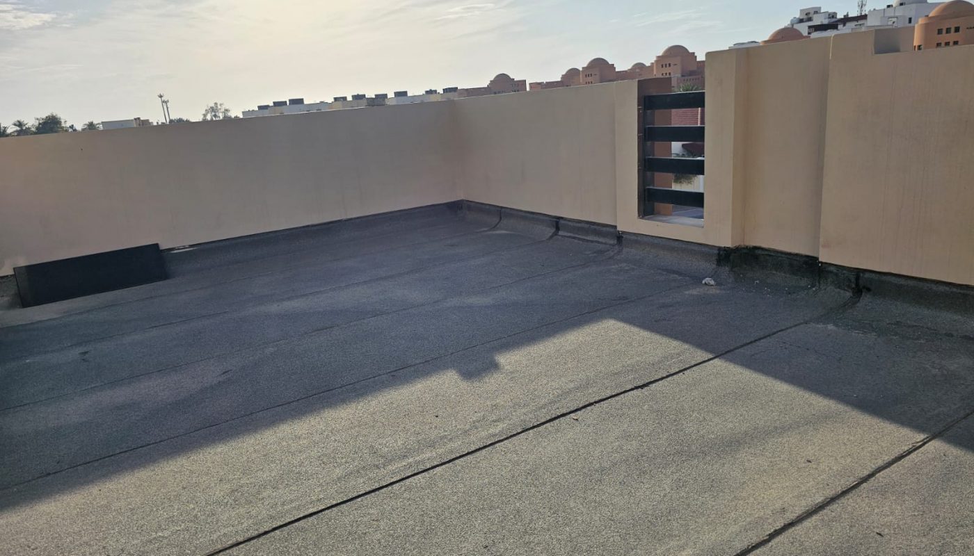 Flat rooftop with concrete surface and low beige walls, partially shaded, under a bright sky with some clouds. Buildings are visible in the background.