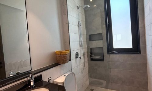 Luxury apartment bathroom with a sink, toilet, shower, and a large mirror, featuring gray tiles and a small window letting in natural light.