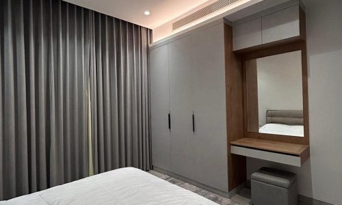 A modern bedroom in a luxury apartment with a bed covered by a white duvet, grey curtains, and wooden furniture including a built-in wardrobe and desk.