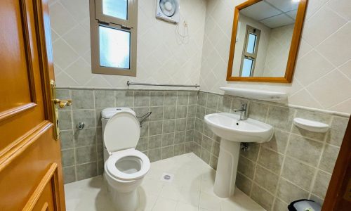 A clean, well-lit bathroom in a furnished apartment with beige tiles, featuring a toilet, pedestal sink, mirror, and frosted windows.