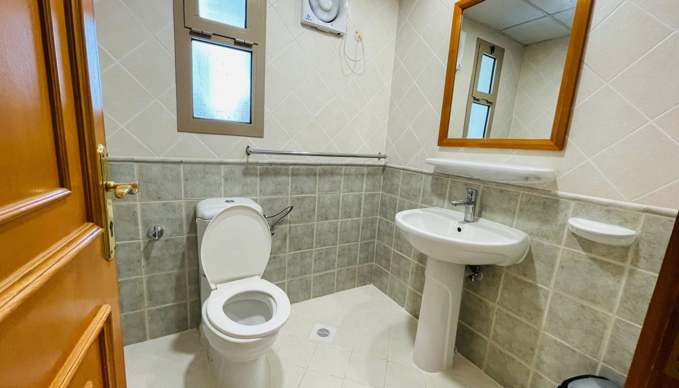 A clean, well-lit bathroom in a furnished apartment with beige tiles, featuring a toilet, pedestal sink, mirror, and frosted windows.