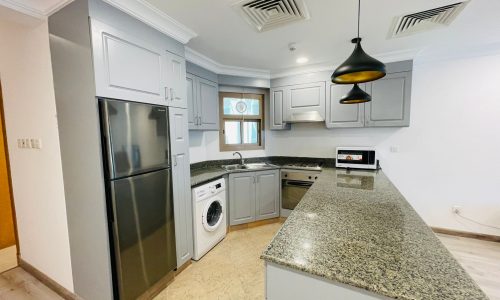 Modern kitchen interior with gray cabinets, stainless steel appliances, a washing machine, granite countertops, and pendant lighting in a furnished apartment.