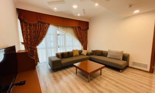 A well-lit living room in a furnished apartment featuring a gray sectional sofa, wooden coffee table, ornate red curtains, and a white ceiling with recessed lighting.