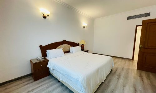 A spacious, minimally furnished bedroom with a double bed, white bedding, wooden nightstand, and wall-mounted lights in the Sanabis area.