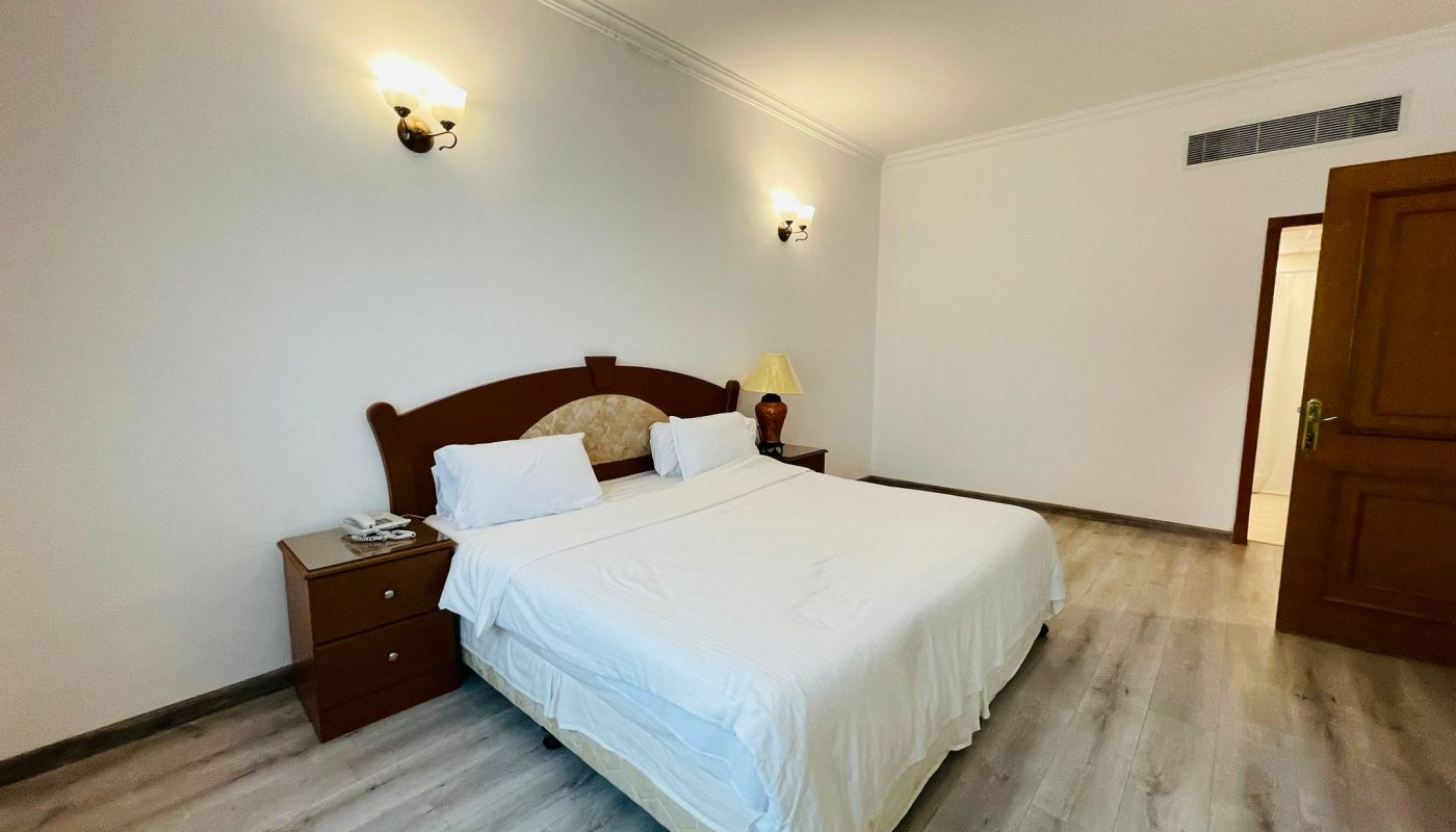 A spacious, minimally furnished bedroom with a double bed, white bedding, wooden nightstand, and wall-mounted lights in the Sanabis area.