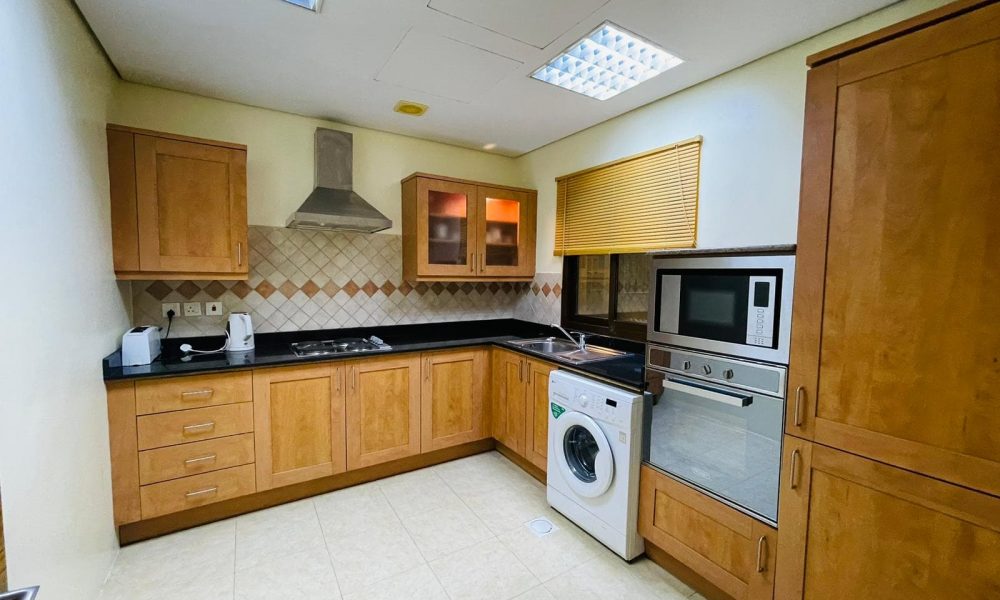 A modern kitchen with wooden cabinets, built-in appliances, and a washing machine under a countertop in a furnished apartment.