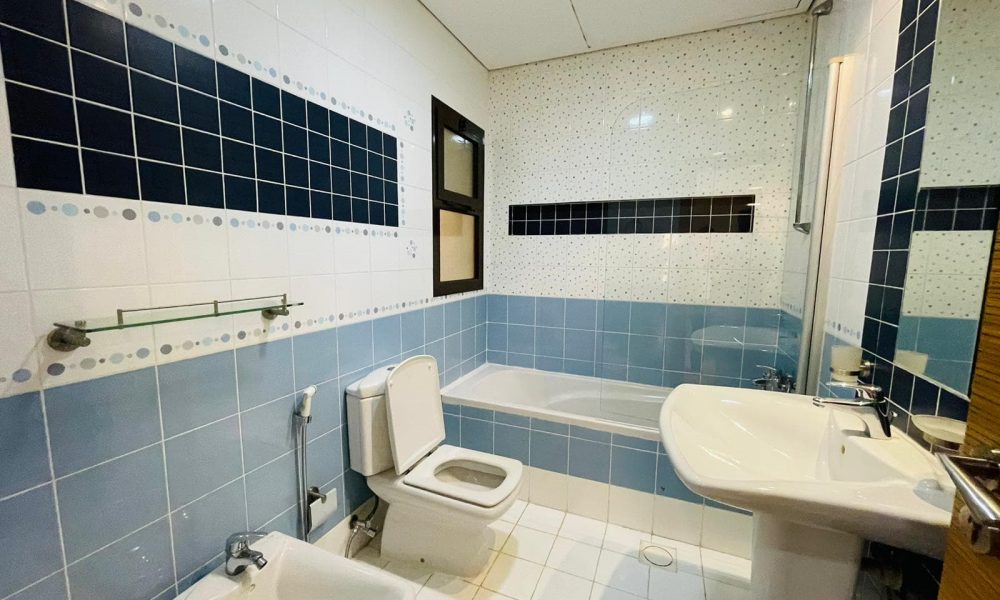 A well-lit bathroom in a furnished apartment for rent in the Sanabis area, with white and blue tiles, featuring a toilet, bidet, bathtub, sink, and a small window.