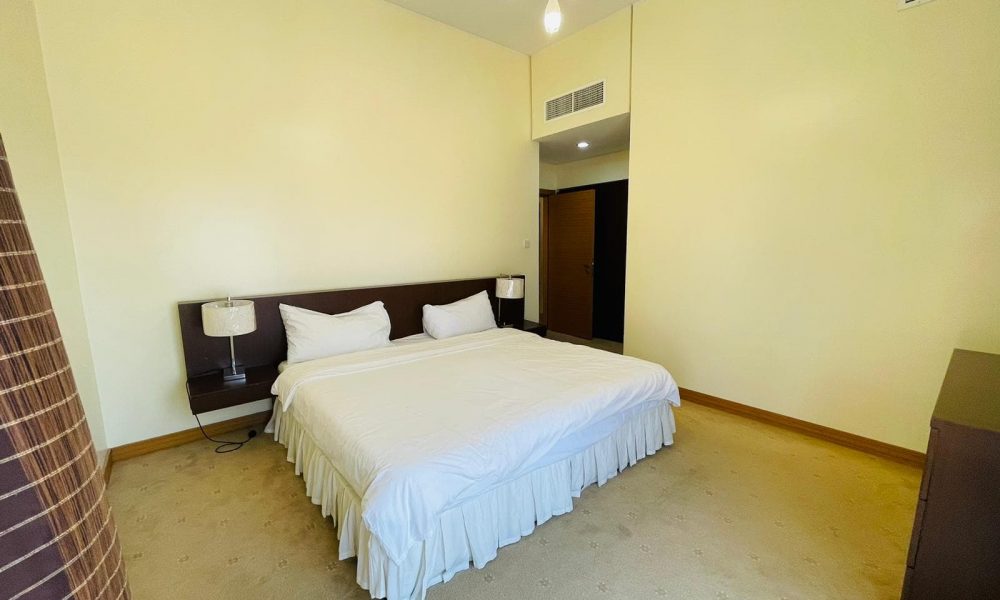 Modern minimalist bedroom in a furnished apartment with a large double bed, white bedding, two bedside lamps, and creamy yellow walls.