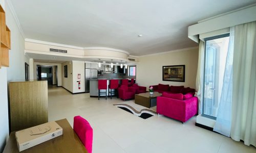 Spacious living room with white tile flooring, vibrant pink sofas, luxury apartment kitchen in the background, and ample natural light from glass doors leading to a balcony.