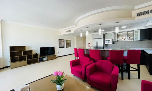 Luxury apartment with a modern living room featuring red sofas, a coffee table with a flower vase, a kitchen bar, and an entertainment unit.