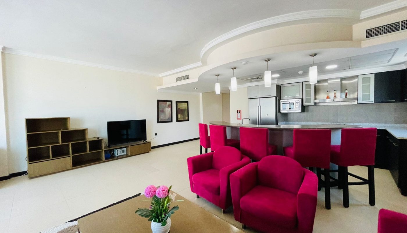 Luxury apartment with a modern living room featuring red sofas, a coffee table with a flower vase, a kitchen bar, and an entertainment unit.