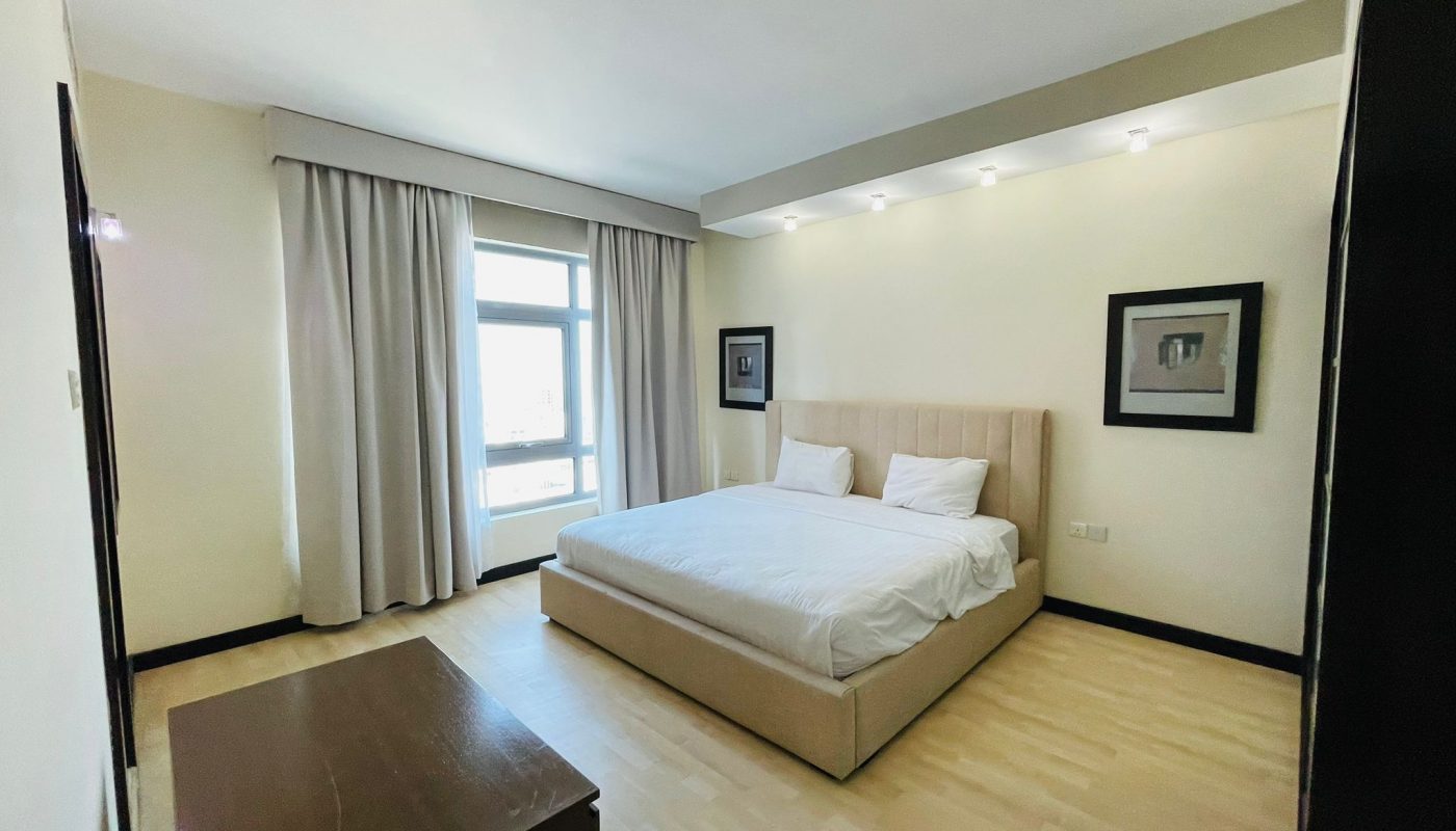 A modern 3 BR apartment bedroom with a large bed, beige headboard, white bedding, and long cream curtains covering a window.