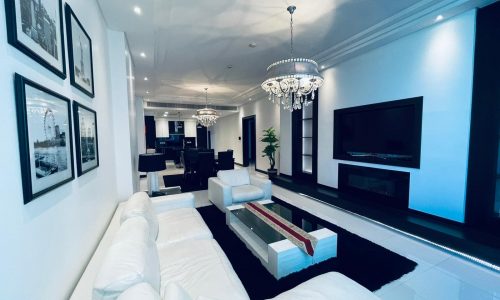 Elegant living room in a luxury apartment with white sofas, a chandelier, and framed pictures on the walls, extending into a dining area.