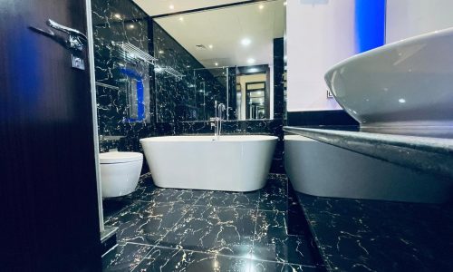 Luxury 2BR Apartment with a modern bathroom, showcasing black marble walls and floor, complemented by a freestanding tub, sink, and toilet. The space is beautifully illuminated by blue accent