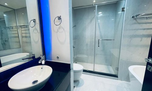 Luxury 2BR apartment bathroom with white fixtures, including a toilet, sink, and shower, accented by blue LED lighting and reflective surfaces.