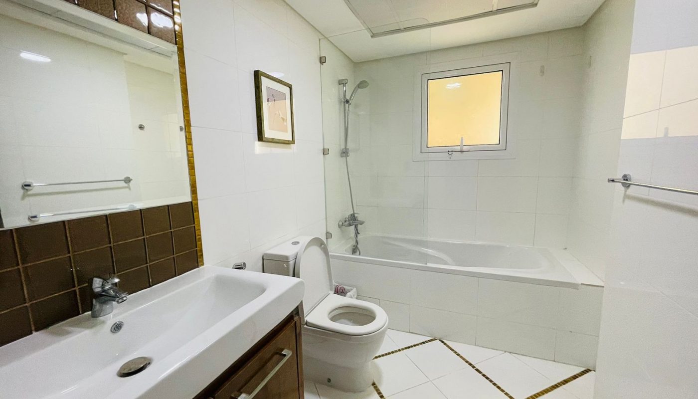 A clean, white tiled bathroom in a luxury apartment featuring a bathtub, toilet, sink, and two framed pictures on the wall, illuminated by natural light from a small window.