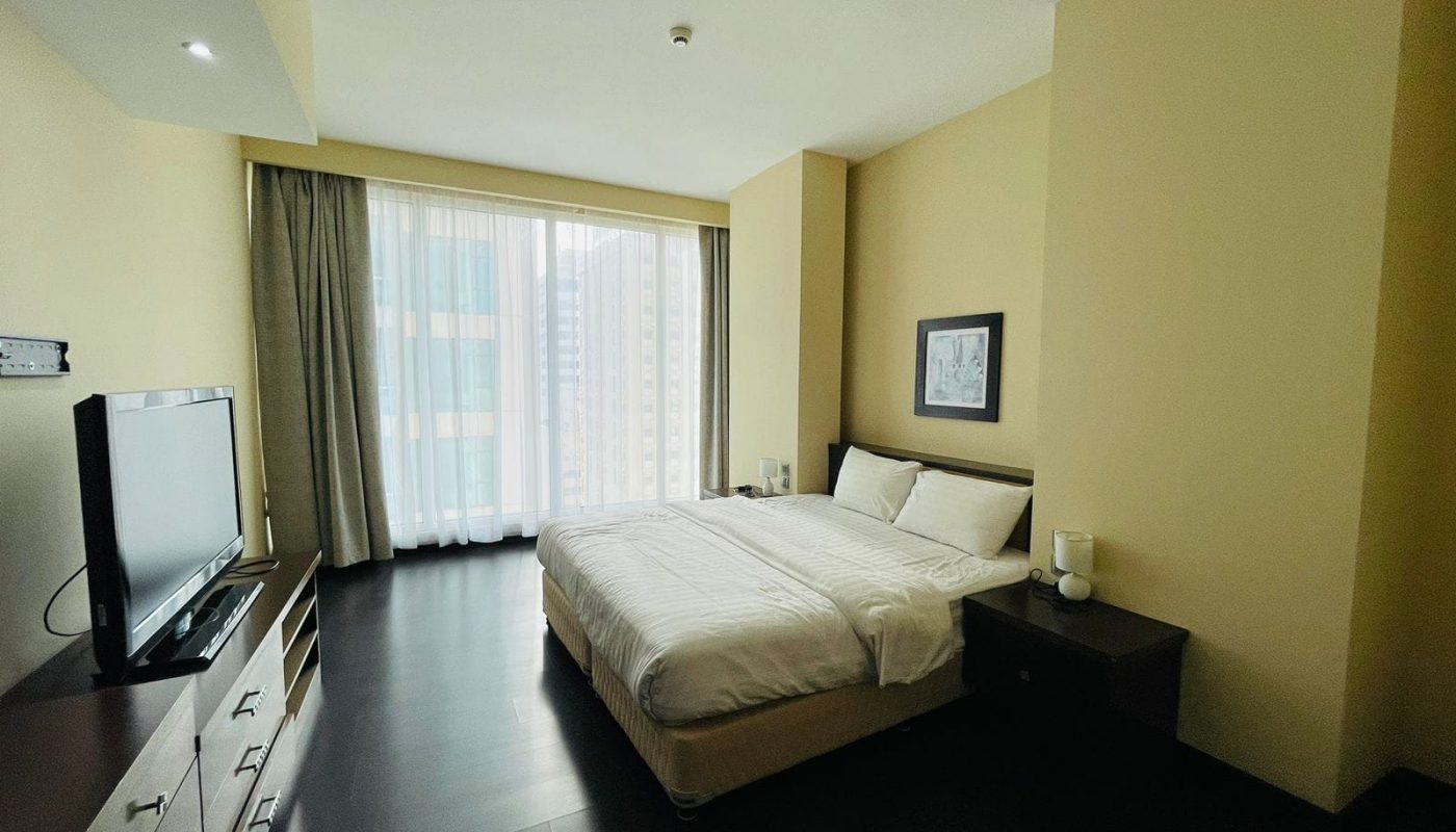 A luxury apartment featuring a modern bedroom with a large bed, beige walls, a dark wooden floor, tv, and a window covered with sheer curtains.