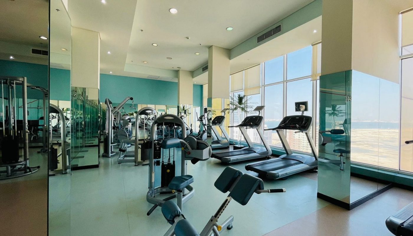 Modern gym interior within a luxury 2BR apartment, featuring treadmills, exercise bikes, and weight-training equipment, with large windows allowing natural light.